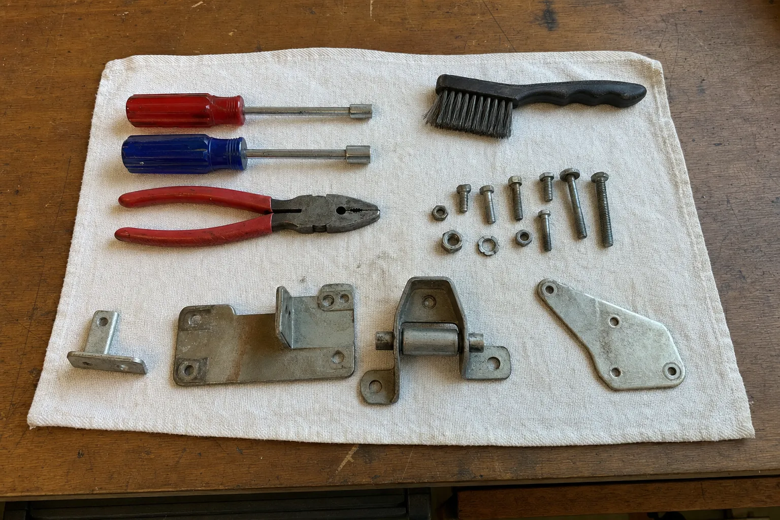 Red and blue nut drivers, lineman pliers and a wire brush on a white cloth beside a small hinge with its pin, two mounting plates, an L-bracket and rows of bolts, nuts and washers